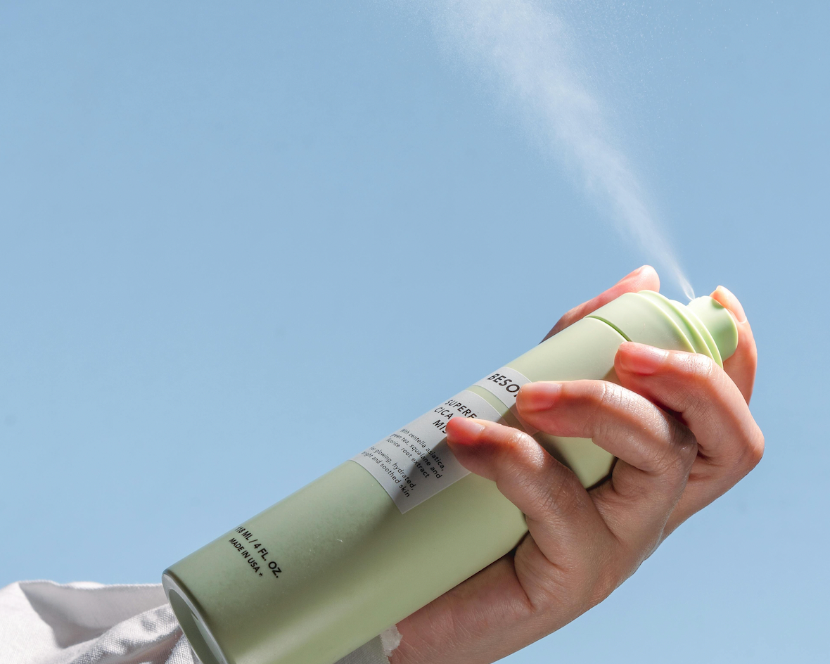 female hand holding a sage green deodorant spraying it with a blue sky aesthetic backdrop
