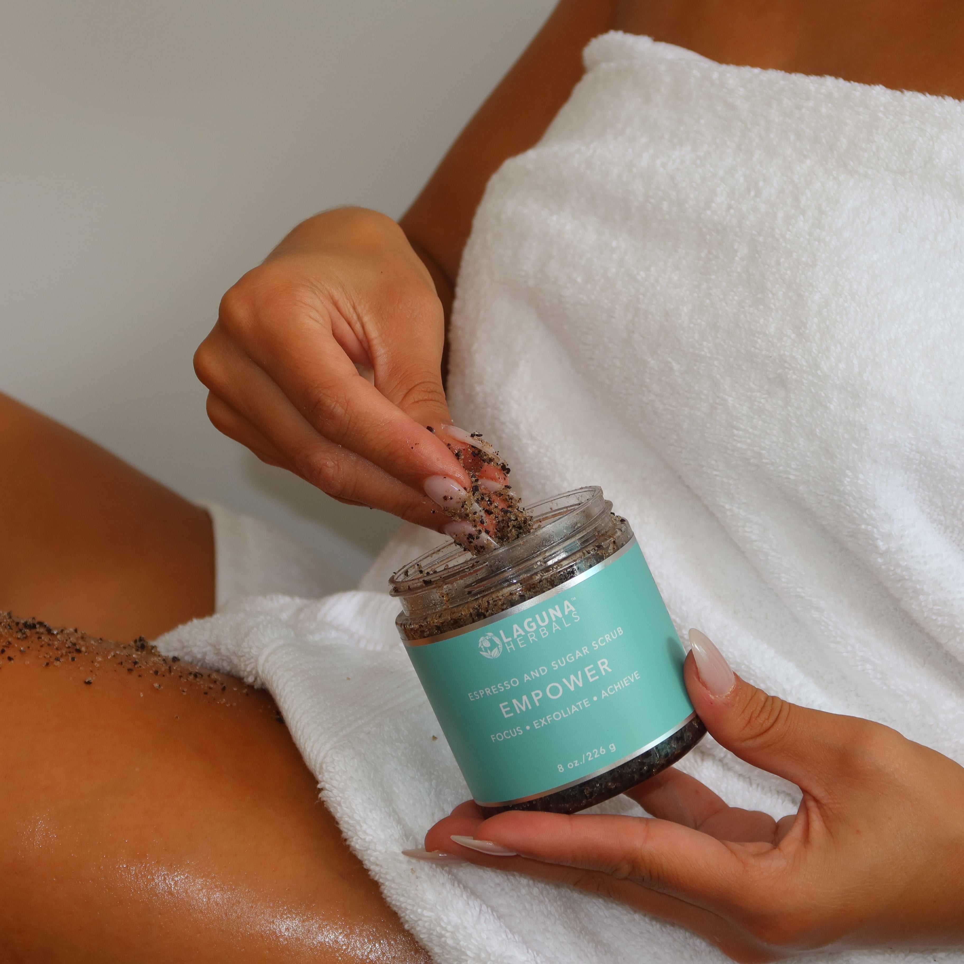 Person applying a exfoliating scrub from a jar labeled 'Body Empower' on their leg.