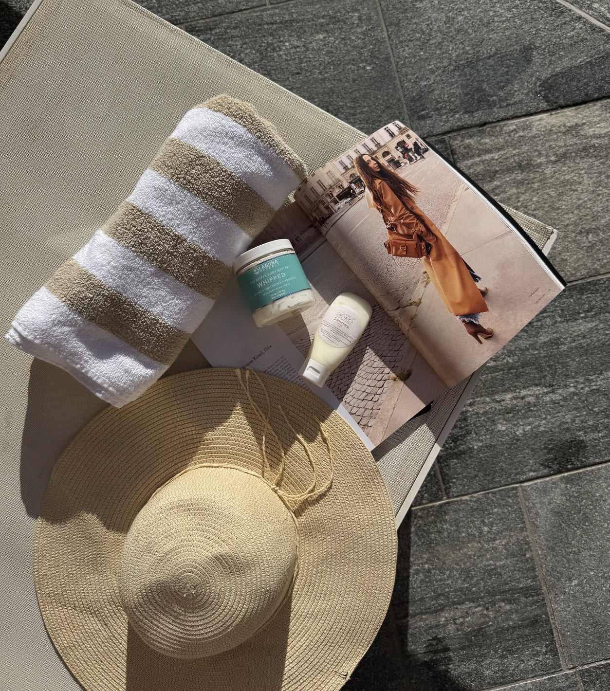 birds eye view image of beach summer essentials with towel, sun hat, summer magazine, sunscreen and body butter