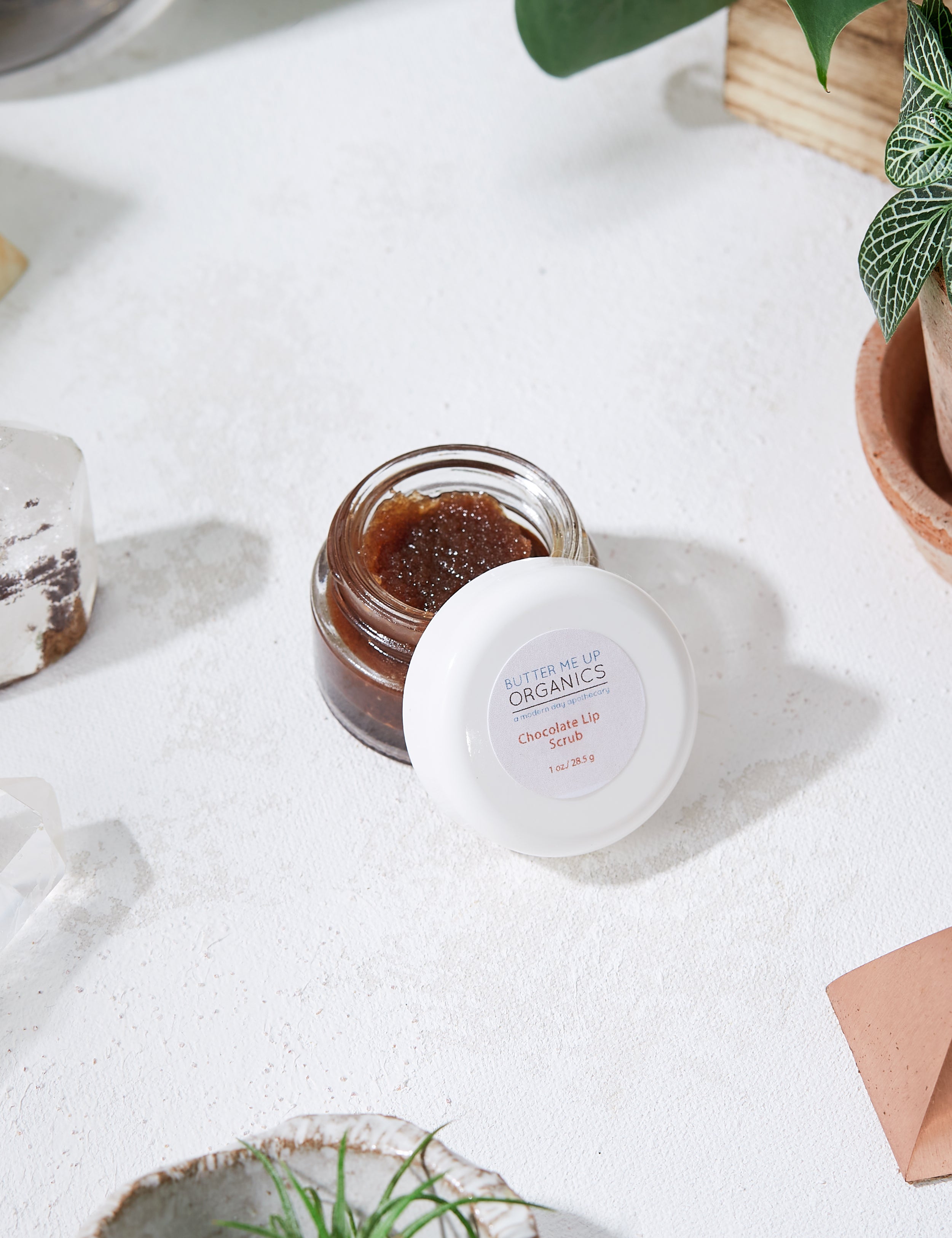 opened product image of the organic Chocolate Lip Scrub with aesthetic white marble backdrop and green leaves