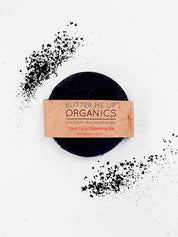 birds eye view of Organic Charcoal Purifying Face Soap Bar product image close up in brown aesthetic packaging but also showing the face bar in an aesthetic white plain backdrop with charcoal sprinkles