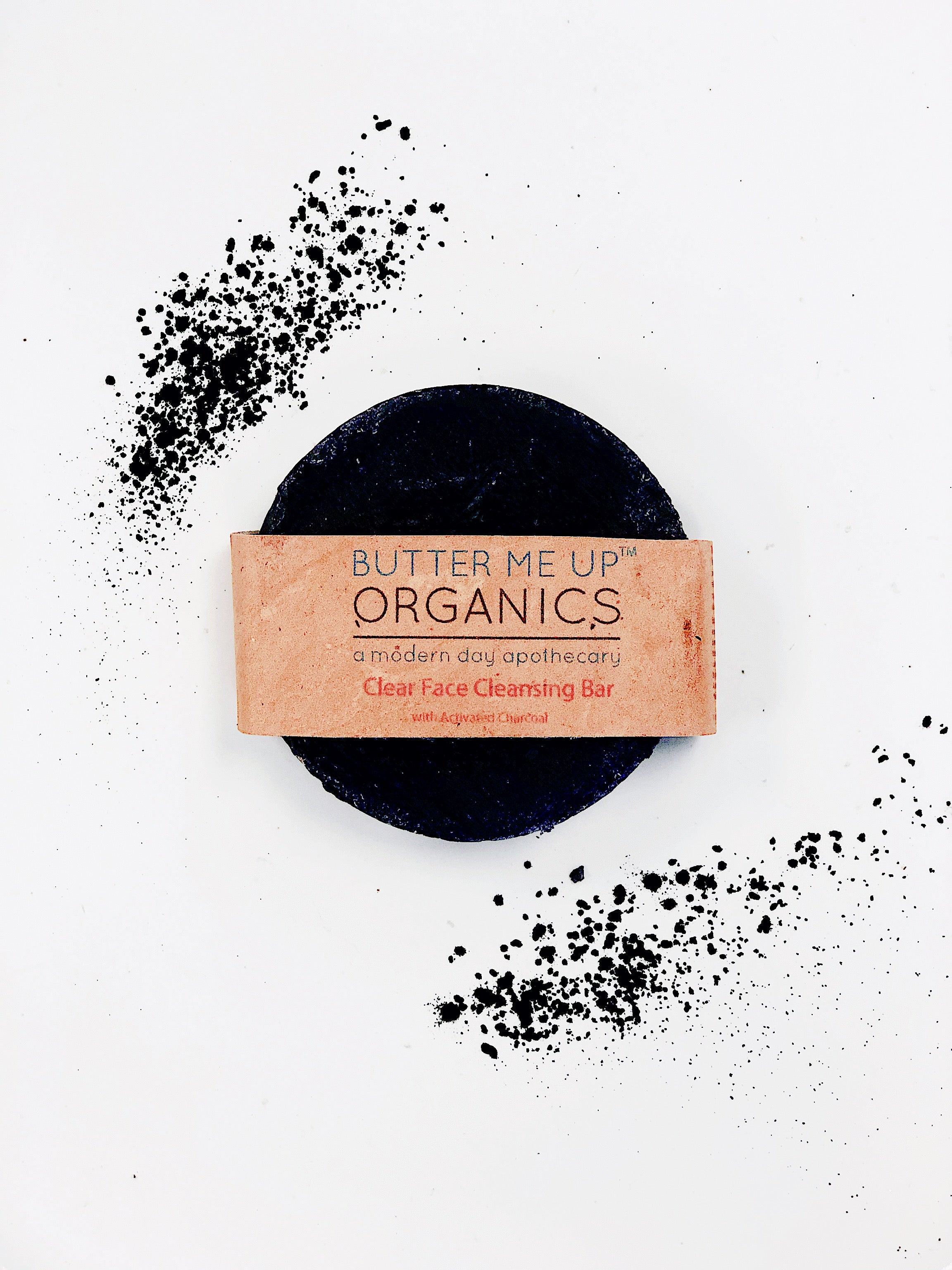 birds eye view of Organic Charcoal Purifying Face Soap Bar product image close up in brown aesthetic packaging but also showing the face bar in an aesthetic white plain backdrop with charcoal sprinkles